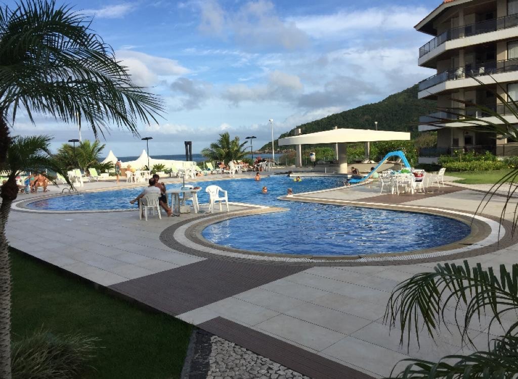 An inviting pool area is surrounded by palm trees, featuring two swimming pools with varying depths. Lounge chairs are positioned around the pools, and a water slide is visible in the background. The area is complemented by shaded seating and a view of the ocean.