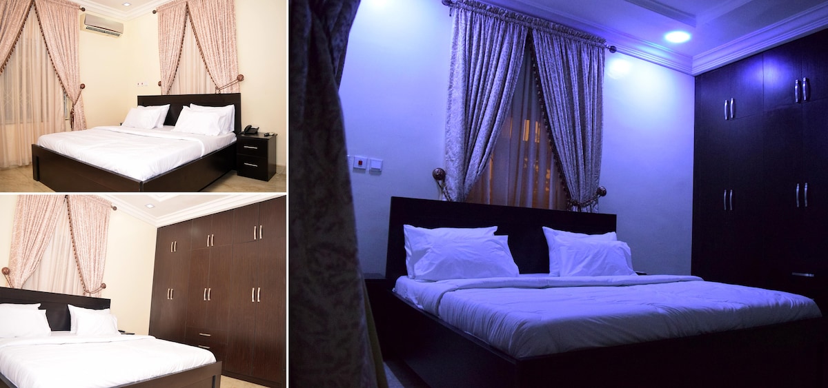 A spacious bedroom features a large bed with white linens and decorative pillows. Soft lighting highlights the room's elegant curtains and dark wooden wardrobes. Natural light filters through the sheer curtains, creating a warm atmosphere.