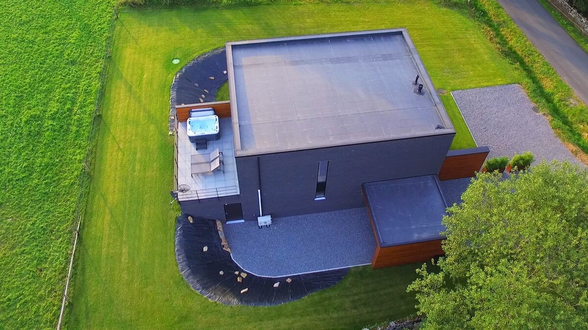 An aerial view of a contemporary building surrounded by lush green grass and a gravel pathway. A private terrace with a heated jacuzzi is visible on the roof. The minimalist design blends naturally with the serene landscape.