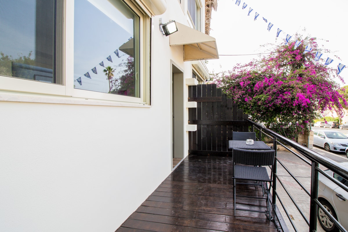 A private terrace is presented with dark wood flooring and a small dining setup. A round table with two chairs is accompanied by flowering pink bougainvillea, while decorative bunting adds a festive touch. A sleek door leads into the apartment.