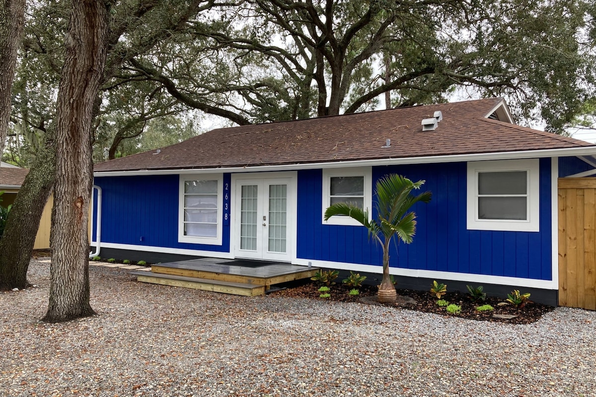 A vibrant blue exterior of the home is highlighted by white trim and large windows. The entrance features a wooden deck with a small step, while surrounding landscaping includes gravel and small plants. Mature trees provide shade in the front yard.
