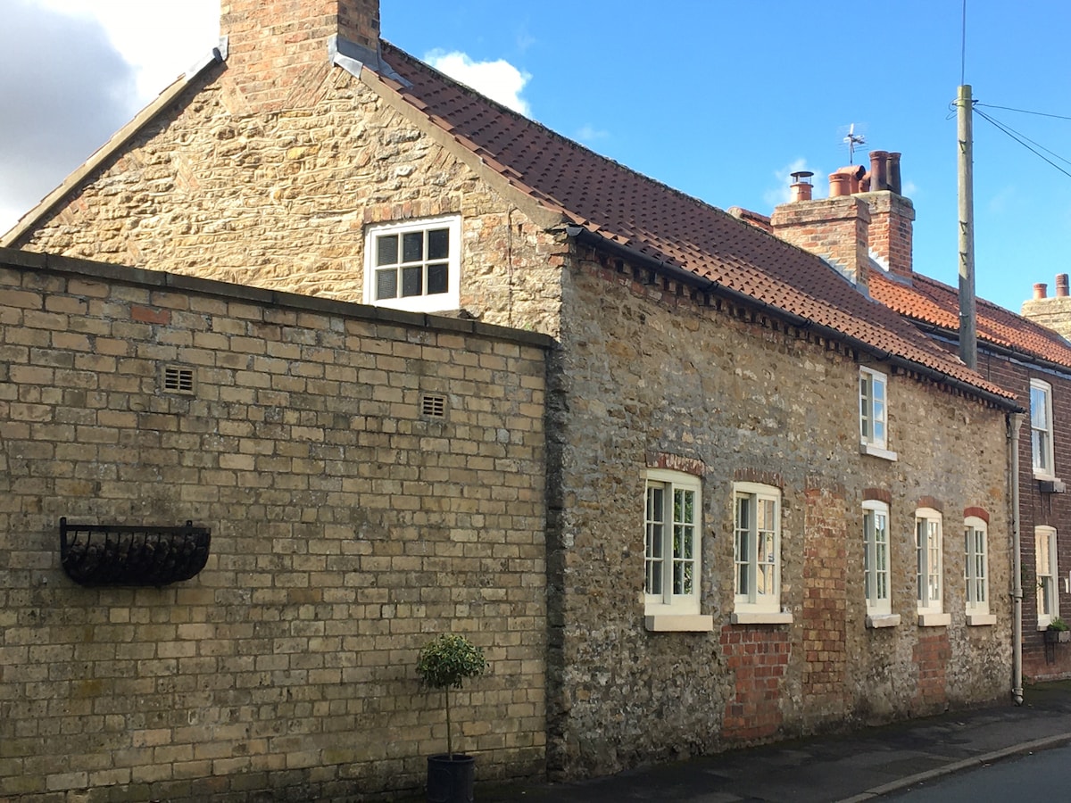 The exterior of the Apple Loft cottage is presented, showcasing its charming stone walls and terracotta roof. Light-colored window frames are featured, alongside a planter with greenery near the entrance. The surrounding village street and sky are visible, contributing to the inviting atmosphere.