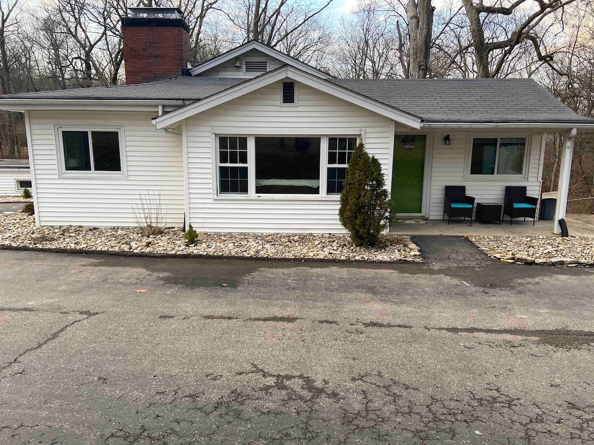 The cottage exterior is presented with a white siding facade and a welcoming green door. Two windows allow ample natural light, and a small porch is adorned with seating. Landscaping features subtle stone accents, enhancing the natural surroundings of trees in the background.