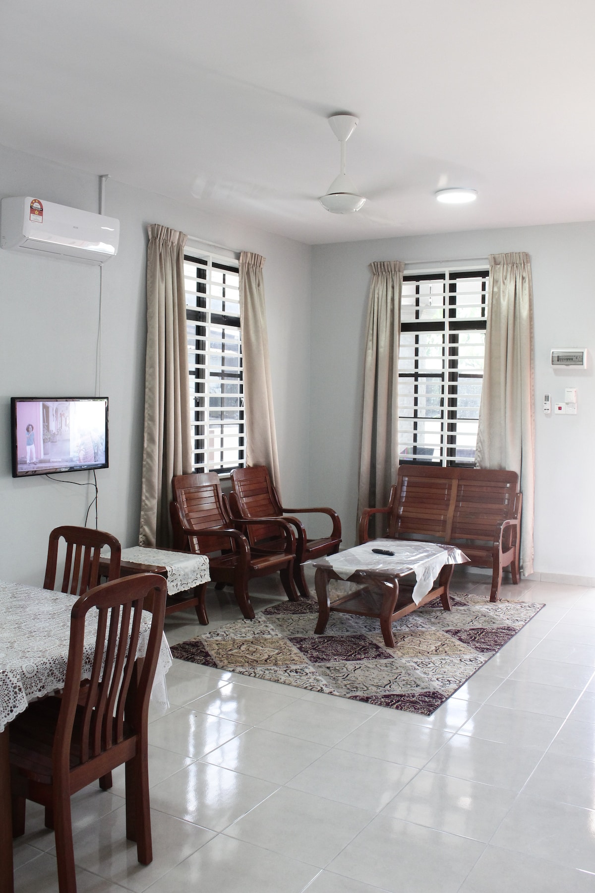 A living area features wooden seating arrangements, including two armchairs and a small table. Light-colored curtains frame windows that allow natural light to fill the space. A television is mounted on the wall, and a small dining table is present, accompanied by chairs.
