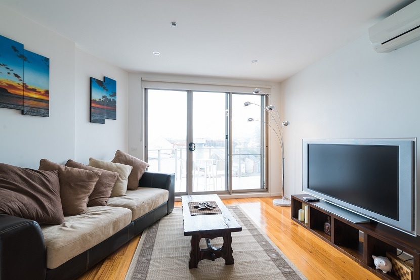 A comfortable living area features a dark leather sofa adorned with plush cushions, complemented by a wooden coffee table and a large TV stand. Natural light enters through sliding glass doors that open to a balcony, while two decorative wall art pieces enhance the ambiance.