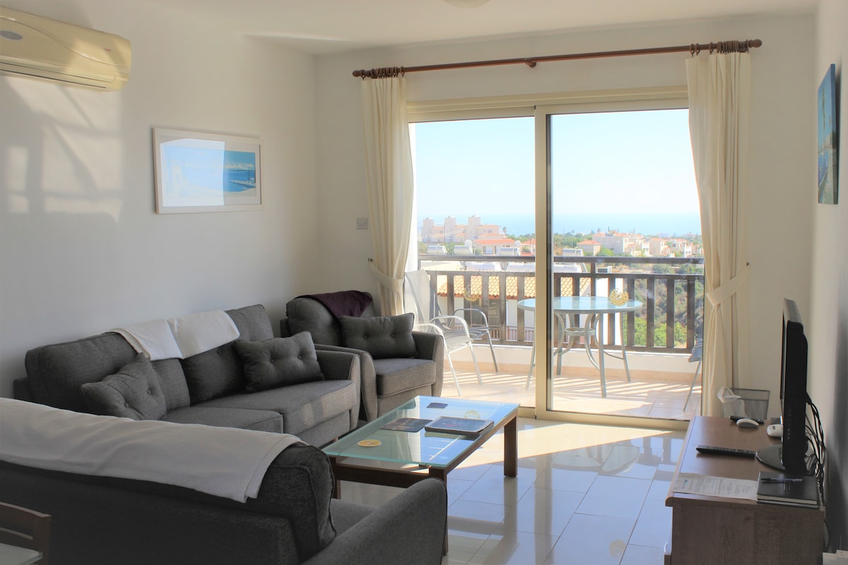 A light-filled lounge area features two sofas and a glass coffee table, with a view through patio doors. The space includes a flat-screen television and decorative artwork. Sunlight illuminates the room, enhancing the inviting ambiance with a backdrop of distant sea views.
