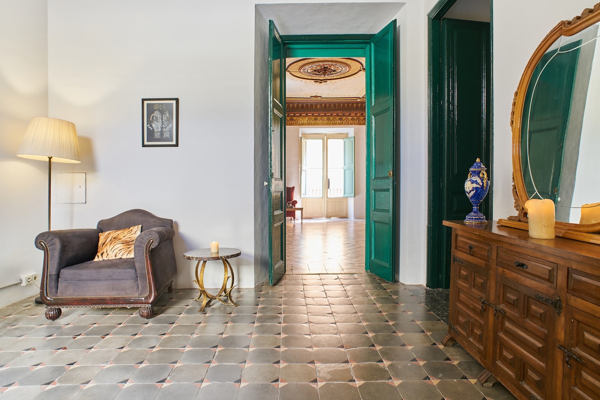 An open area is displayed with elegant green doors leading into a spacious room. A cozy armchair with a patterned cushion sits beside a small side table. The tiled floor features a classic pattern, and natural light illuminates the space, highlighting the decorative details of the interior.