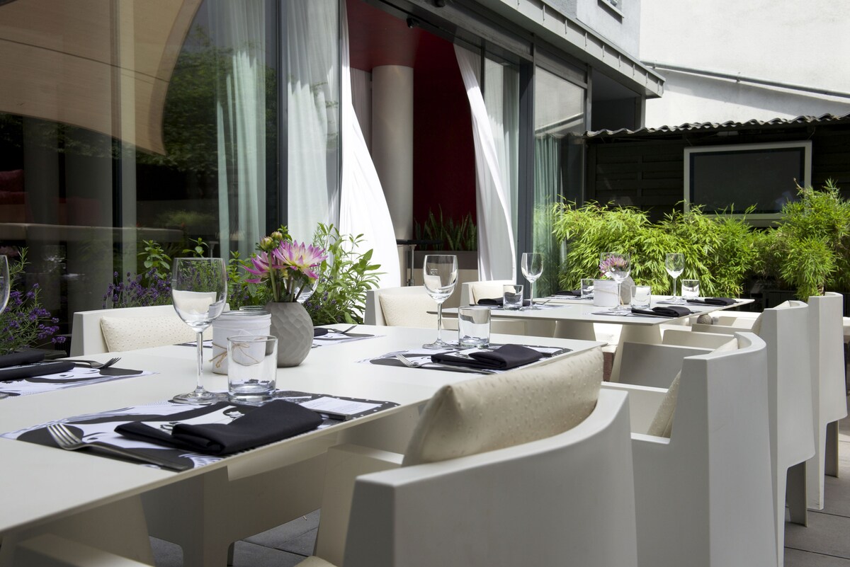 An outdoor dining area is presented, featuring a white table surrounded by modern chairs. Elegant glassware and dark napkins are neatly placed on the table, complemented by lush greenery in planters nearby. Sheer curtains gently frame the space, enhancing a tranquil atmosphere.