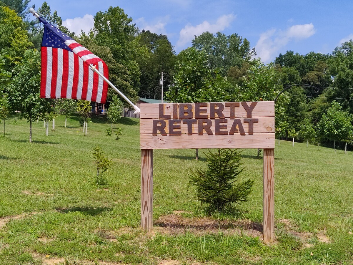 The wooden sign for Liberty Retreat is prominently displayed, featuring bold lettering against a clear blue sky. An American flag flutters nearby, and the surrounding grassy area is dotted with young trees, contributing to the rural and welcoming ambiance.