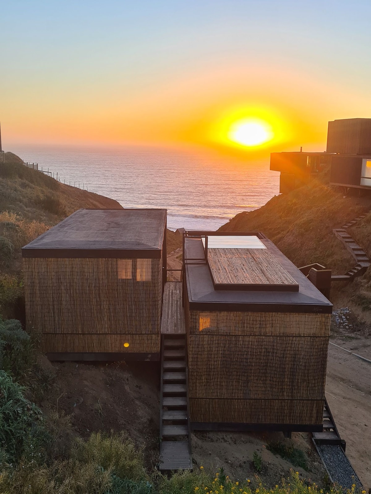 Two cabins are positioned on a sloping landscape, framed by the sea and a vibrant sunset in the background. The cabins feature wooden exteriors and are connected by a set of steps leading down toward the coastline.