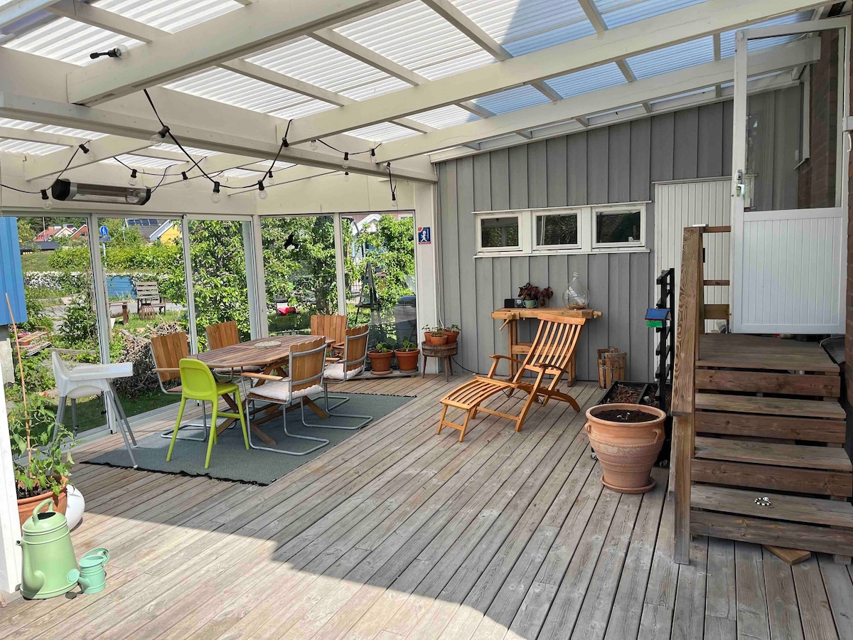 A spacious patio area features a wooden deck, surrounded by greenery and potted plants. A large dining table with several colorful chairs is positioned alongside lounge chairs. Natural light filters in through a roof structure, creating an inviting outdoor space.