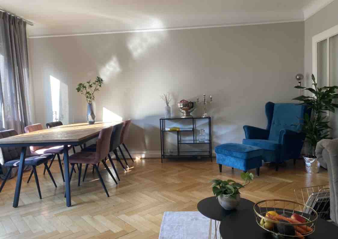 A spacious living and dining area features a large wooden table surrounded by six upholstered chairs. A bright blue chair adds a pop of color, while natural light enters through large windows. A side table holds decorative items, and greenery enhances the relaxed atmosphere.