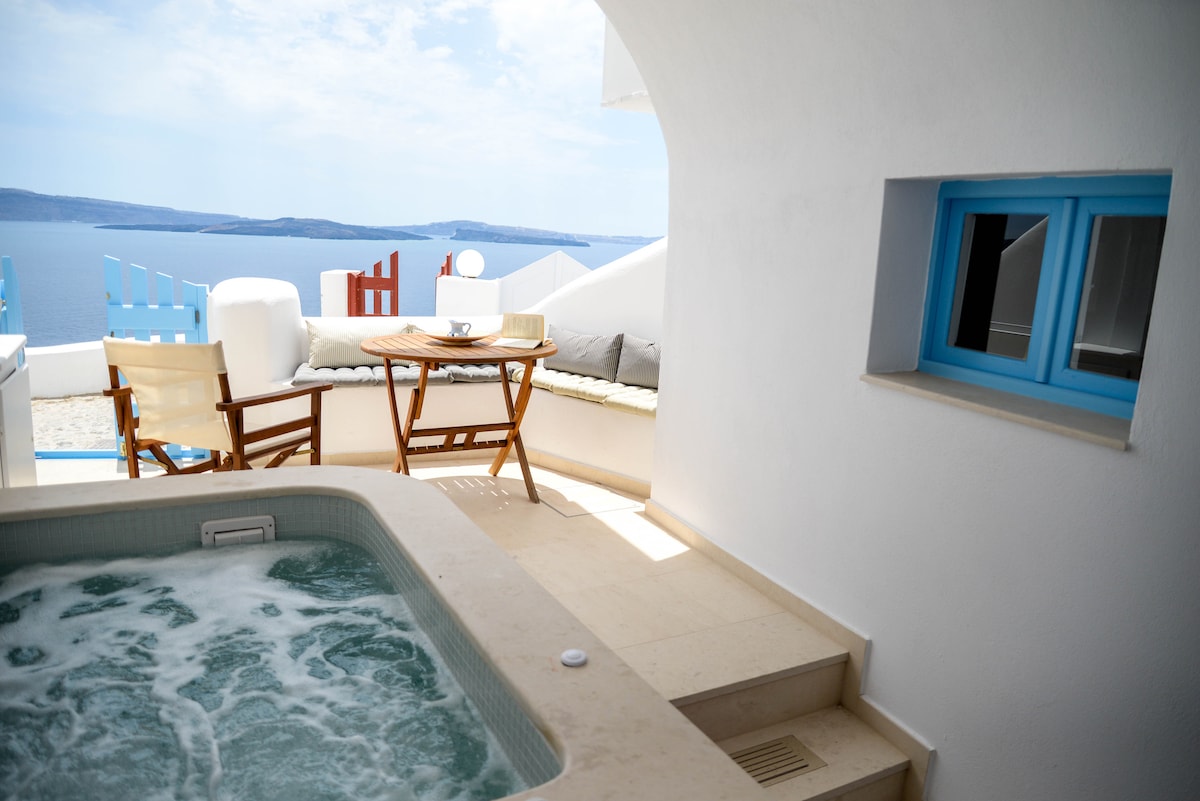 An outdoor area features a heated jacuzzi surrounded by white walls, with a wooden table and two chairs positioned nearby. A cozy seating nook is integrated into the space, with a striking blue window offering a view of the Aegean Sea and sky.