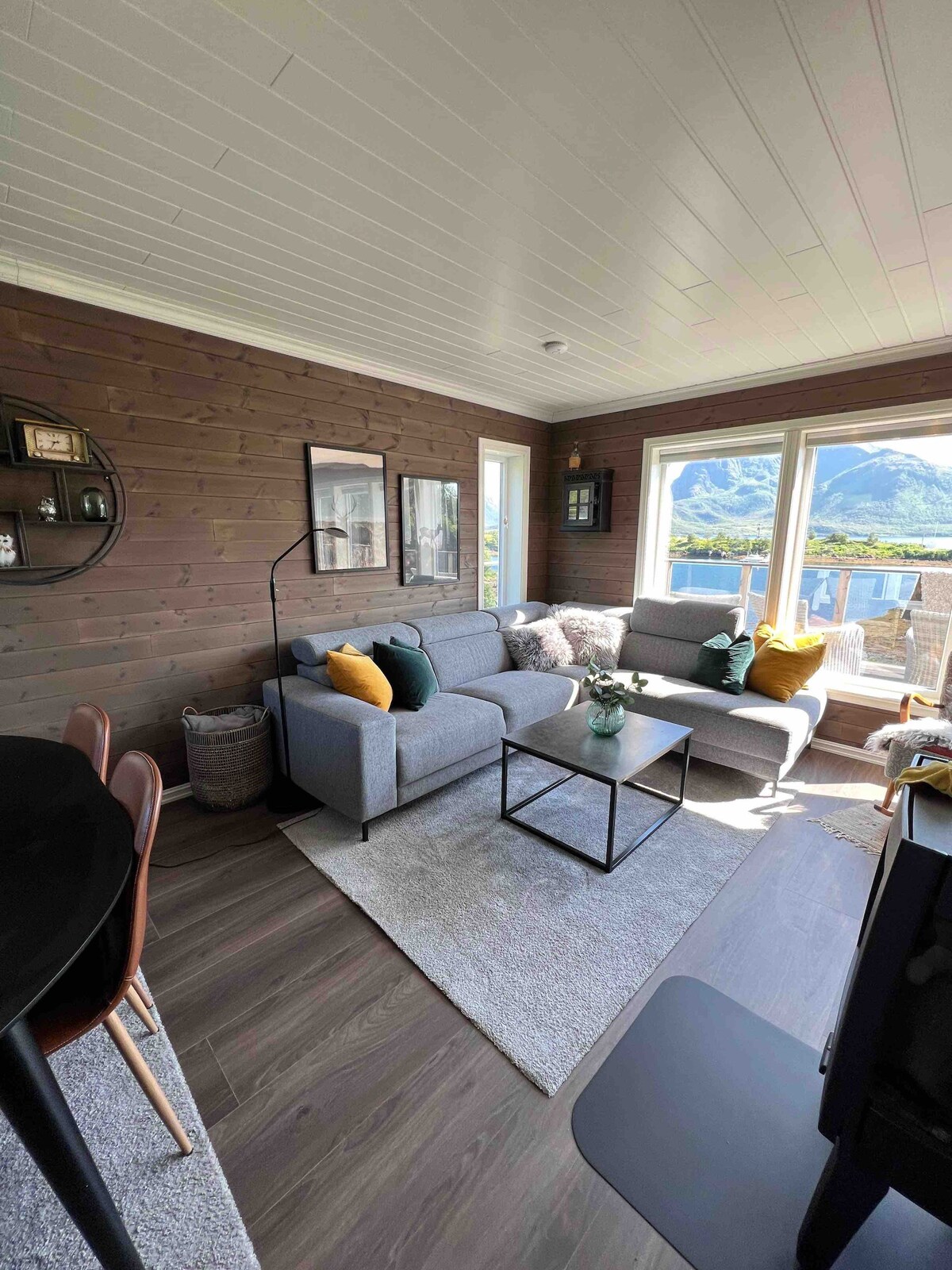 A comfortable living area is displayed, featuring a light grey sectional sofa adorned with cushions in various colors. A black coffee table is centrally positioned on a soft area rug. Large windows offer views of the surrounding mountains and allow natural light to fill the space.