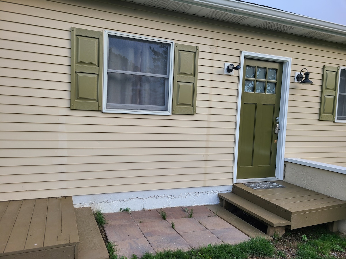 The exterior of a light-colored house is shown, featuring a green door with antique-style knobs. Two green shutters frame a window above the entrance. A small porch with wooden steps leads to a paved pathway, surrounded by grass and greenery.