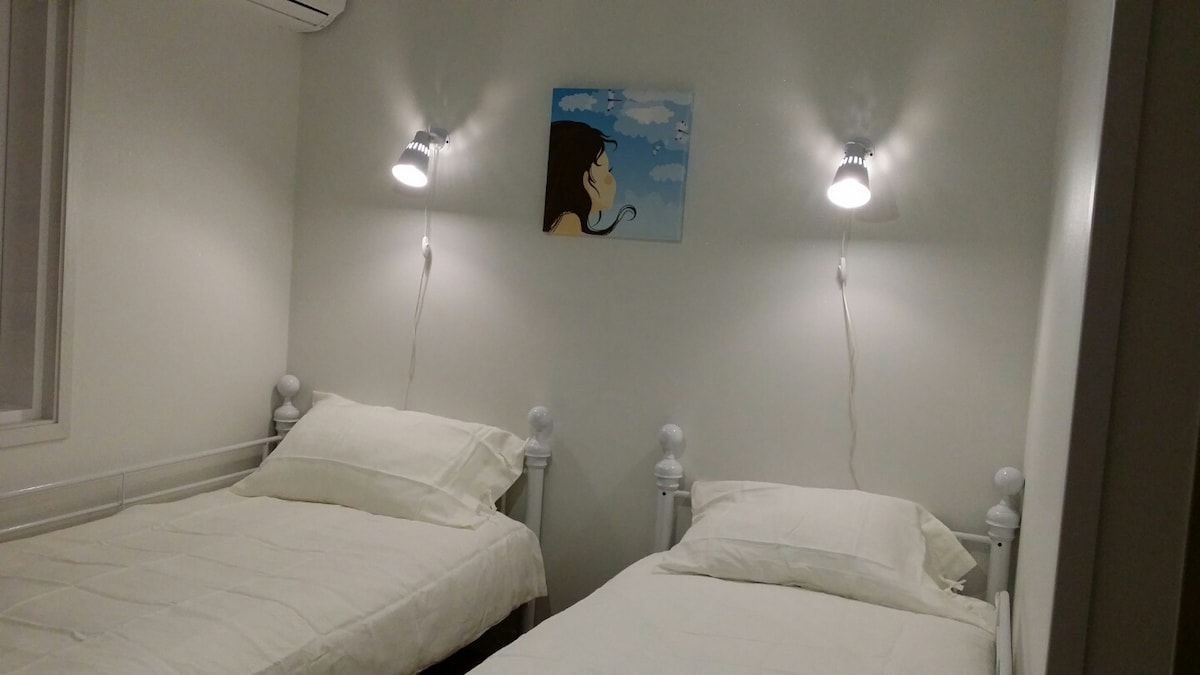 A bedroom features two twin beds with white bedding, positioned against a bright wall. Each bed is paired with a bedside lamp mounted on the wall. A piece of artwork depicting a profile against a blue sky adds a touch of color.