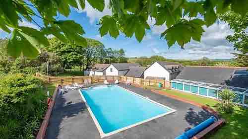 A spacious outdoor heated pool is surrounded by a landscaped area with trees and greenery. Several lounge chairs are positioned around the pool's edge, and buildings can be seen in the background, set amongst the natural landscape of the holiday village.