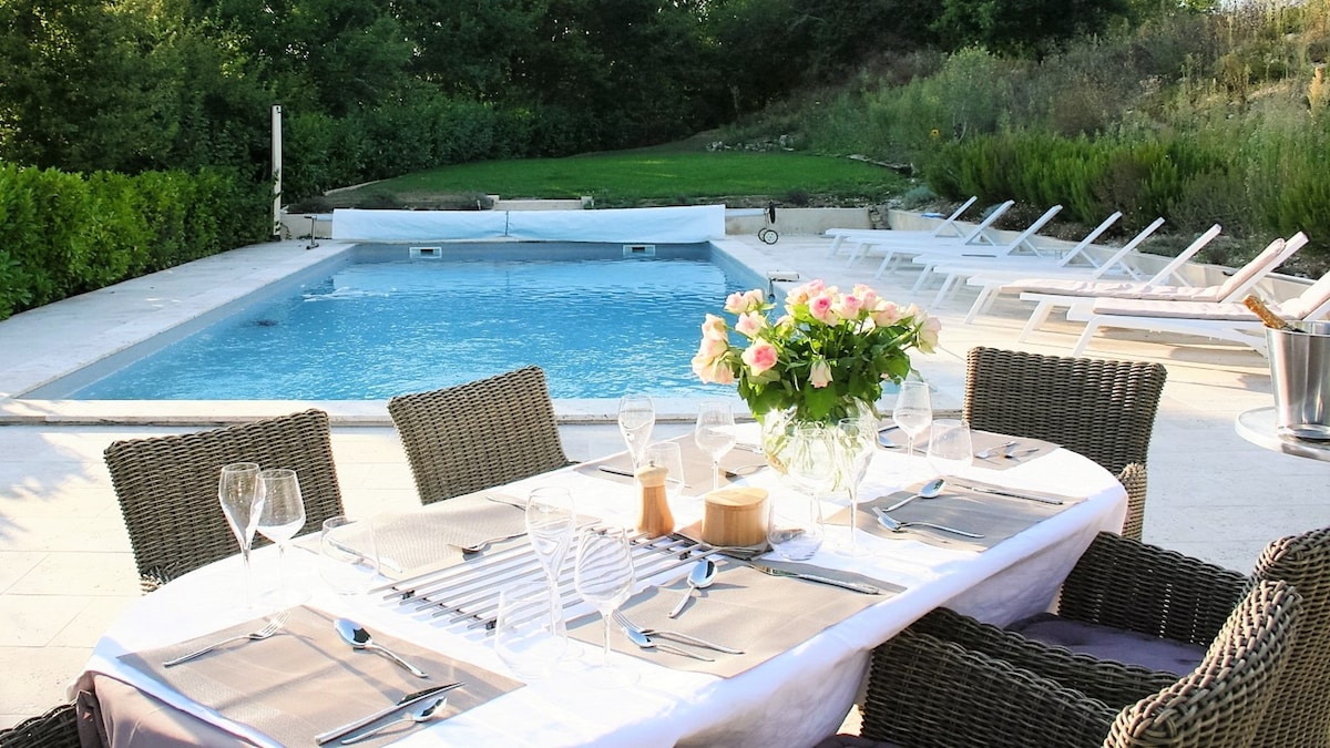 An outdoor dining area is set beside a large, private pool. A table is beautifully arranged with place settings and a bouquet of flowers. Comfortable seating surrounds the table, while numerous sun loungers are positioned near the pool, framed by greenery.