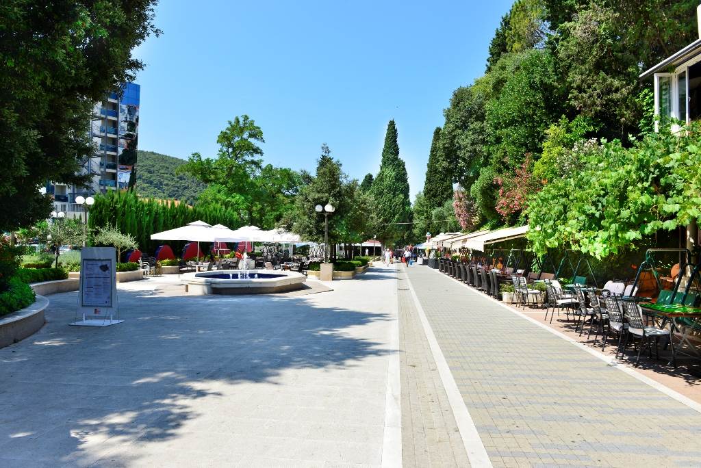 A pedestrian pathway is bordered by lush greenery and trees, leading to dining areas with shaded umbrellas. A fountain is visible along the path, and various seating options are arranged neatly, inviting guests to relax and enjoy the surroundings in the sunshine.