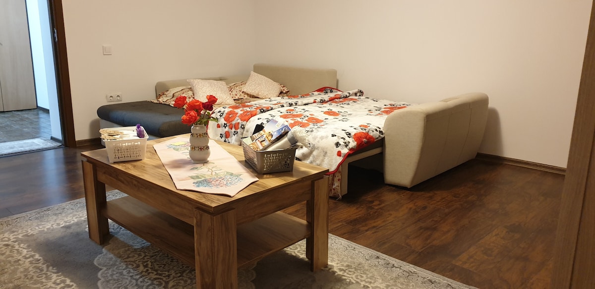 A spacious sleeping area is presented with a large sectional sofa bed, adorned with a colorful floral blanket. A wooden coffee table is positioned in the center, featuring a basket and a vase with fresh flowers. Neutral walls and warm wooden flooring enhance the room's inviting feel.