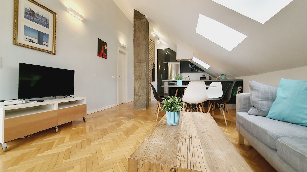 An open living area with wooden flooring features a cozy gray sofa adorned with turquoise cushions. A wooden coffee table and a potted plant are situated in the foreground. Bright light enters through skylights, illuminating a modern kitchen area in the background.