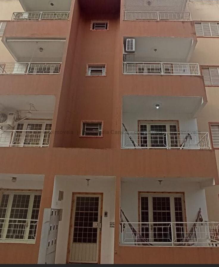 The exterior of a multi-story apartment building is presented, showcasing a warm color scheme and multiple balconies. A central entrance is visible, flanked by evenly spaced windows and air conditioning units. Railings add a decorative element to the balconies.