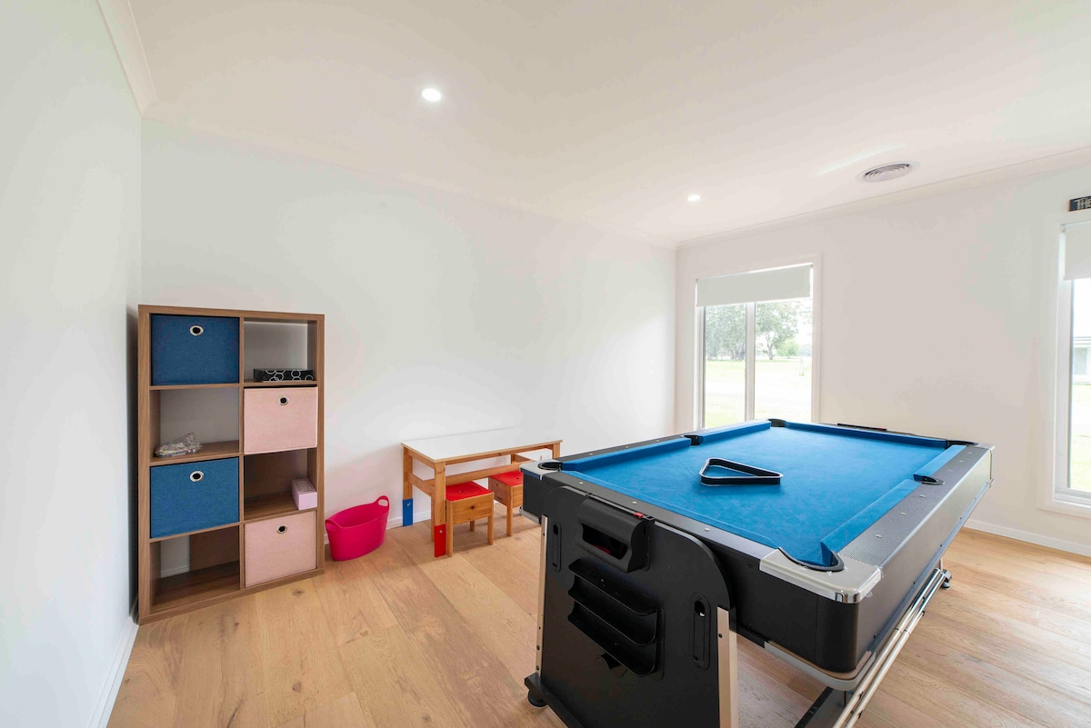 A dedicated recreational room features a blue pool table and a small wooden table with two chairs. A colorful shelf holds storage bins, promoting organized play. Natural light filters through large windows, enhancing the spaciousness of this inviting area.