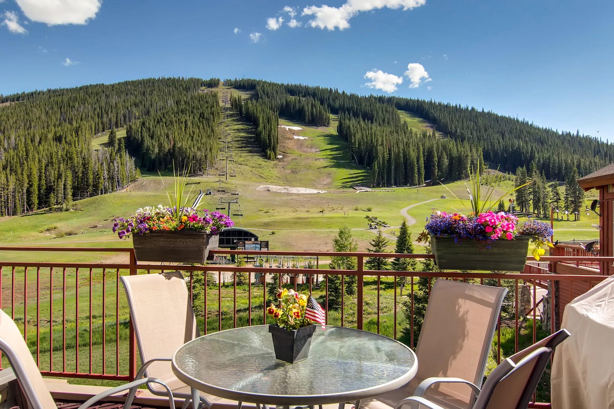 A spacious private deck offers a round glass table surrounded by four chairs. Bright flower pots add color to the scene, with a backdrop of verdant hills leading to ski slopes under a clear blue sky.