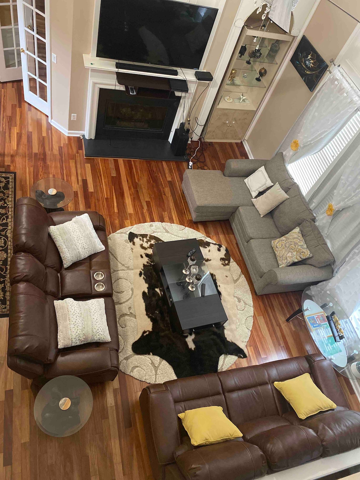 A spacious living area features a mix of seating options, including two leather sofas and a gray sectional, arranged around a stylish coffee table. A large area rug complements the hardwood flooring. Natural light filters through the nearby windows, enhancing the inviting space.