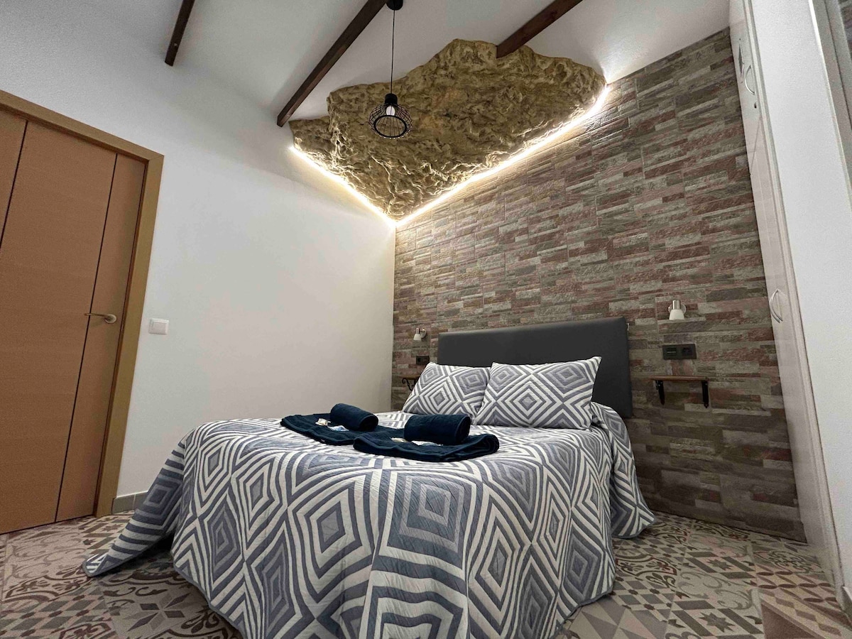 A spacious sleeping area is showcased with a large bed covered in a patterned blanket. A unique illuminated rock feature is seen above, complemented by a textured wall. The room includes modern lighting fixtures and a neutral color palette, creating a calming environment.