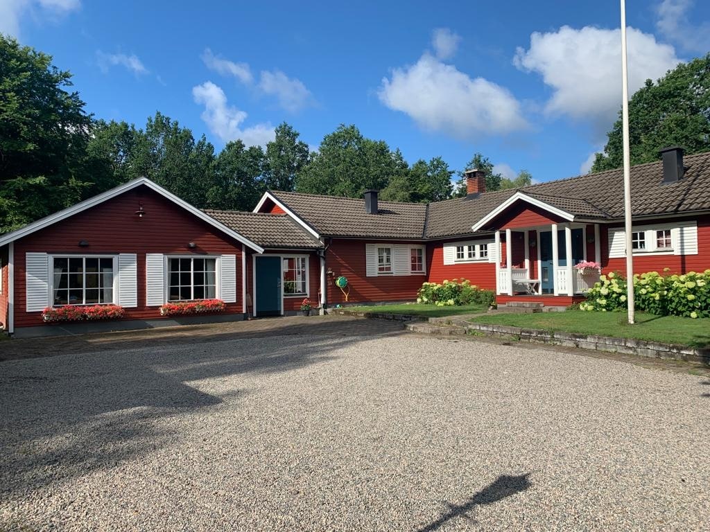 A charming red house with a pitched roof is set on a gravel driveway. Surrounding greenery includes trees and flowering plants, enhancing the natural setting. Large windows reflect natural light, and a welcoming front door provides access to the inviting interior.
