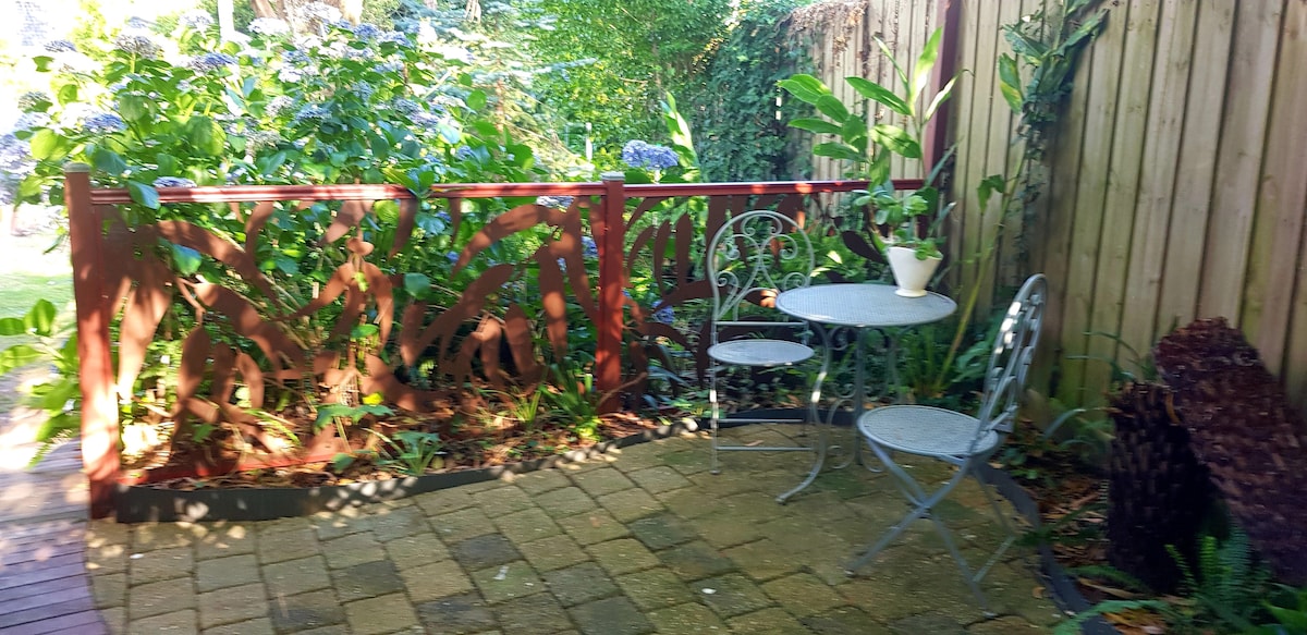 A private outdoor patio features two metal chairs and a small table, surrounded by lush green plants and vibrant flowers. A wooden fence with decorative cutouts offers a sense of enclosure, while the paved area provides a comfortable space for relaxation.