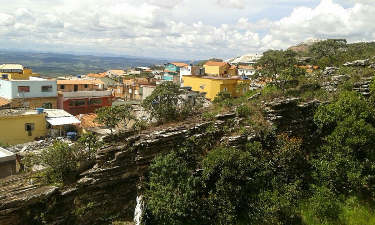 A panoramic view of a town reveals colorful buildings nestled among green vegetation and rocky terrain. The landscape showcases rolling hills under a partly cloudy sky, with the distant horizon presenting a sense of openness and tranquility.