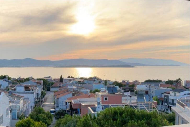 A serene view captures the landscape at sunset, showcasing the rooftops of nearby homes complemented by the glistening sea. Subtle colors blend in the sky, enhancing the tranquil atmosphere. The distant mountains add depth to the beautiful coastal scenery.