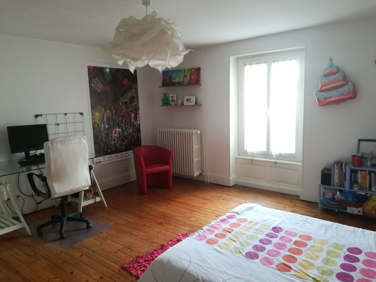 A spacious bedroom features a large bed adorned with a colorful polka-dot bedding set. A work desk and chair are positioned beside a window that allows natural light to enter. Decorative elements, including wall art and a playful light fixture, add character to the room.
