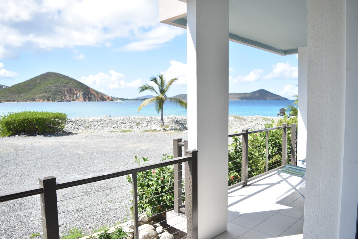 A patio area features a railing with greenery and stunning views of the azure waters and distant mountains. Bright sunlight reflects off the water, creating a serene and inviting ambiance. The space includes a small seating area for relaxation.