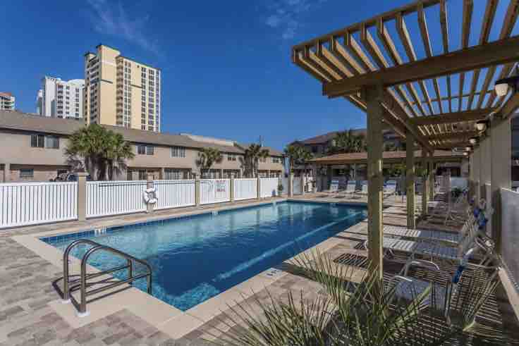The heated pool area features a large swimming pool surrounded by lounge chairs, shaded by pergolas. Lush greenery and a white fence encase the space, while the buildings of the complex are visible in the background under a clear blue sky.