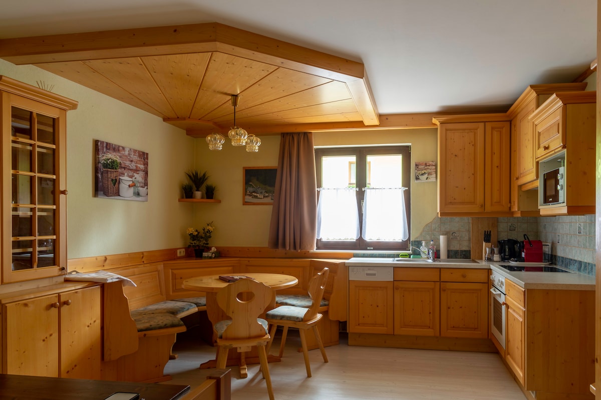 A wooden kitchen features light-colored cabinetry and a circular dining table surrounded by cushioned chairs. Natural light enters through a window, highlighting decorative plants and artwork on the walls. An under-counter stove and microwave are integrated into the space.