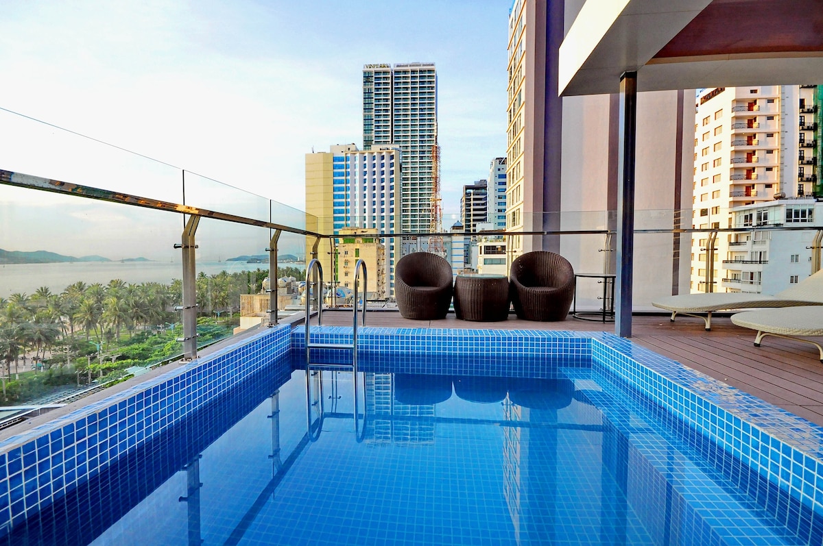 An outdoor pool with blue tiled flooring is situated on a spacious terrace, framed by modern loungers. The distant skyline of Nha Trang with tall buildings and palm trees is visible in the background, offering a sense of urban relaxation.
