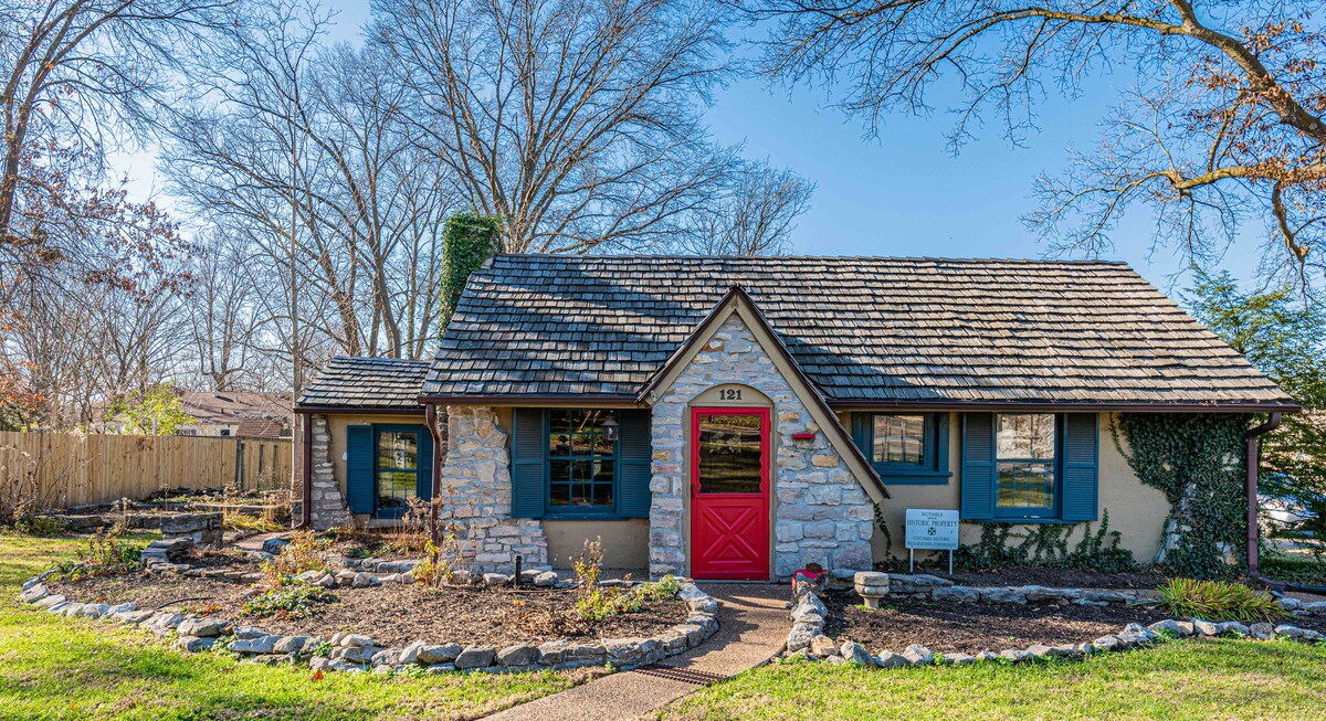 A charming cottage exterior is visible, featuring a stone facade and a distinct red door that adds a pop of color. Surrounding landscaping includes native plants and a stone border, contributing to the inviting curb appeal. Tall trees stand in the background against a clear blue sky.
