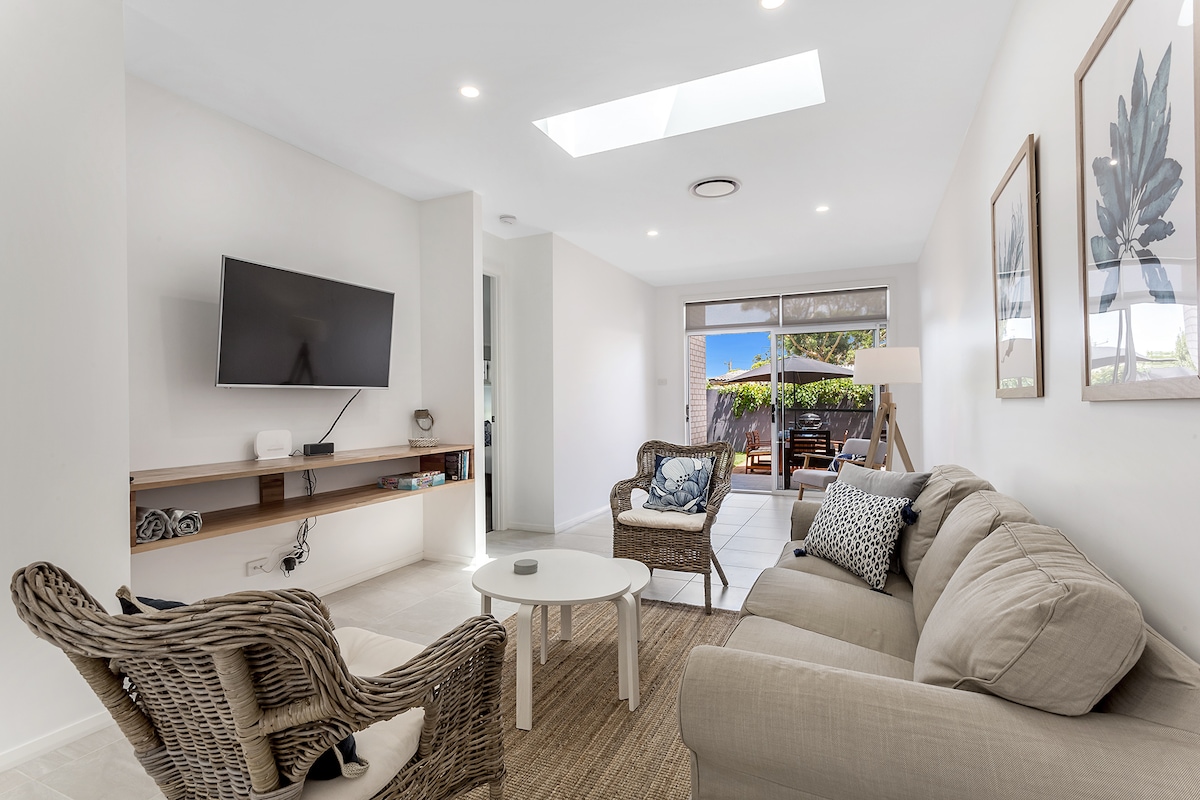 A modern living area features a comfortable sofa and armchairs, accompanied by a light coffee table. Natural light enters through a skylight and glass doors that lead to an outdoor space. A flat-screen TV is mounted on the wall above a wooden shelf.
