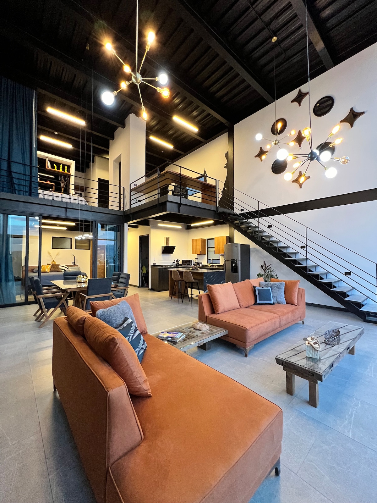 An open-concept living area features two orange sofas arranged around a wooden coffee table. A modern light fixture hangs above, while large windows invite natural light. The staircase leads to an upper level, providing a view of the dining and kitchen space.
