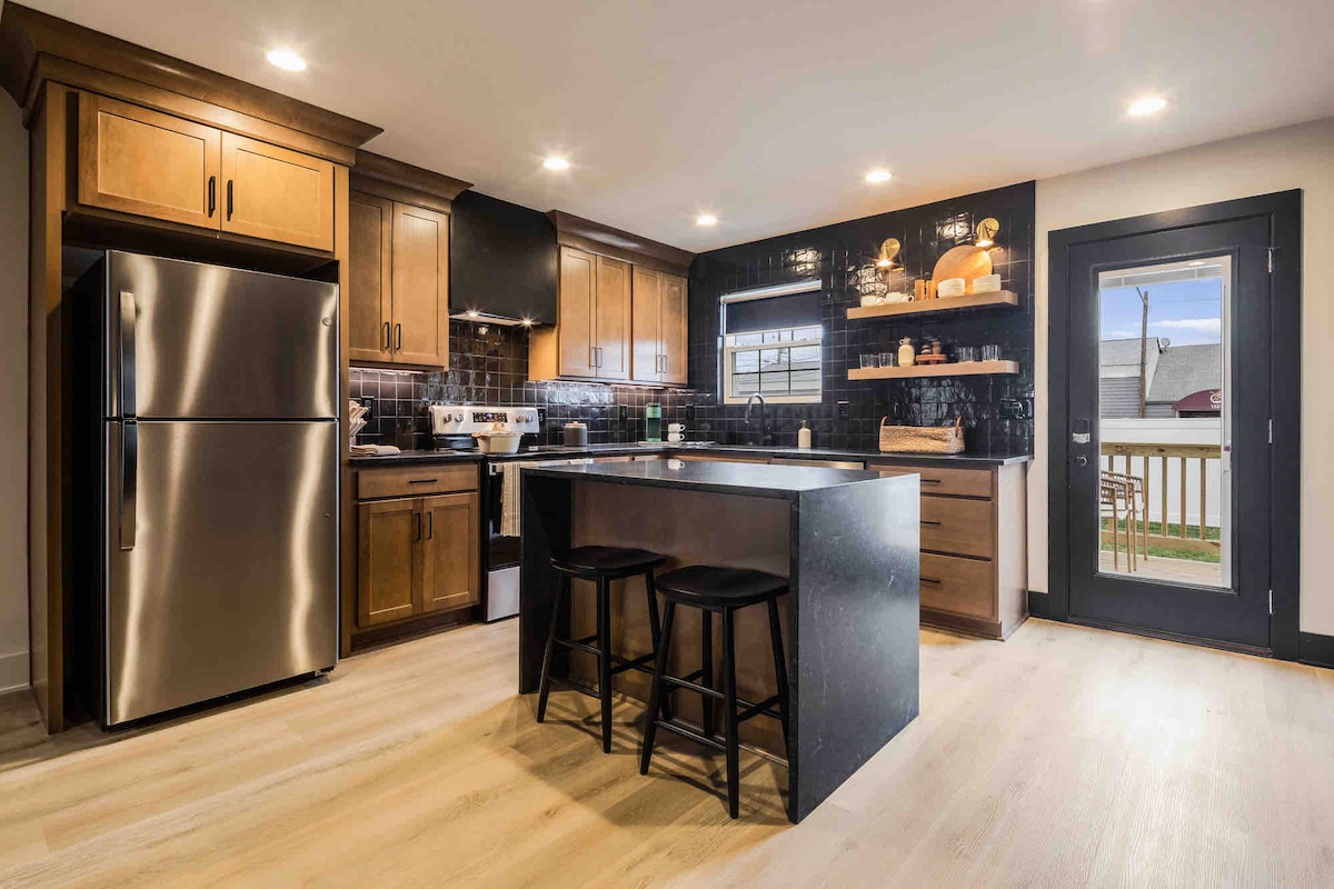 A modern kitchen features wooden cabinetry with a sleek stainless steel refrigerator. A central island with bar stools provides additional preparation space. Natural light enters through the window above the sink, while a door opens to the outdoor area.