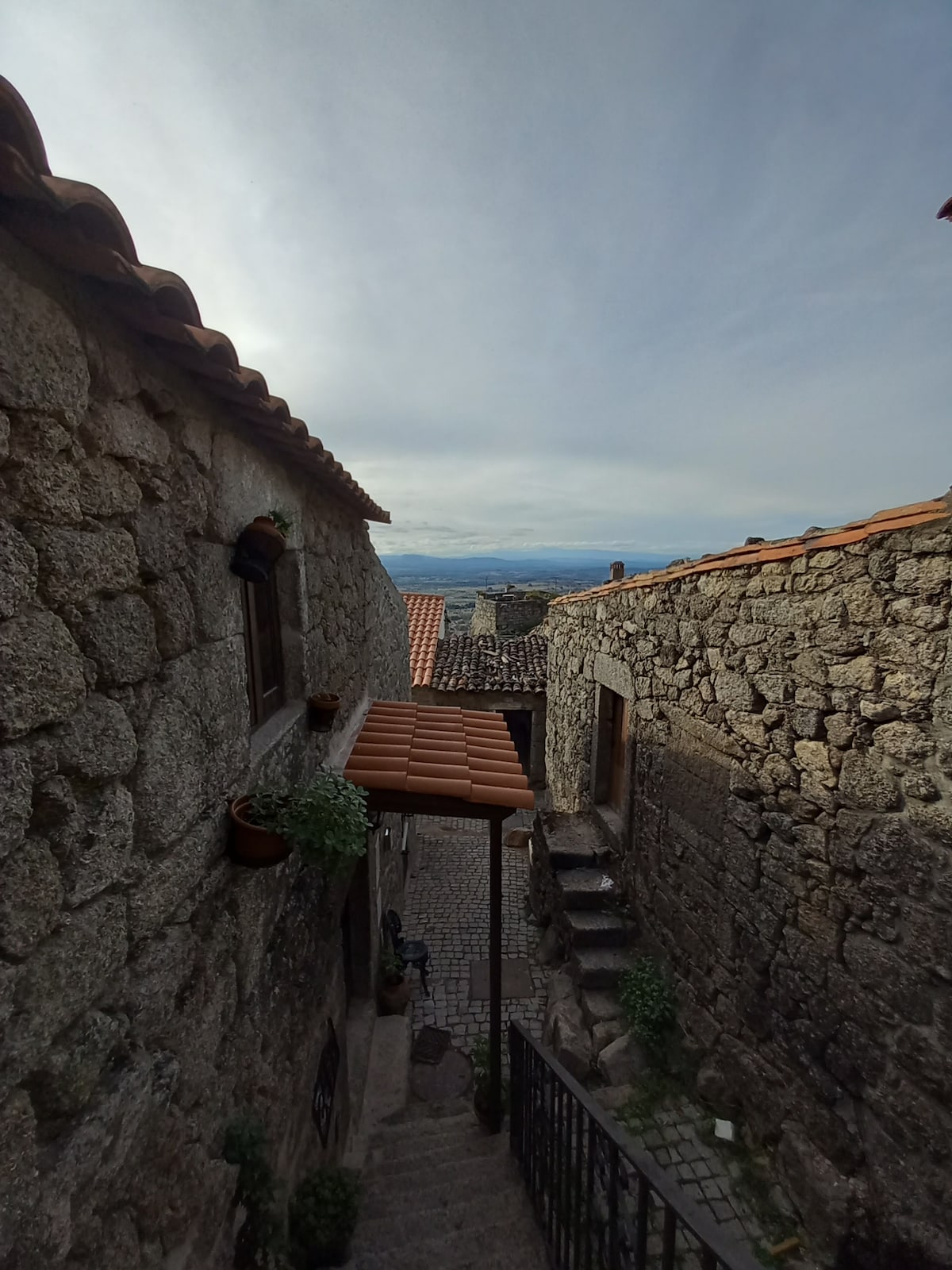 A narrow stone pathway leads through a charming alleyway lined with rustic buildings. Potted plants are placed along the walls, and a glimpse of the distant landscape is visible above the rooftops. The scene captures the medieval essence of the village.