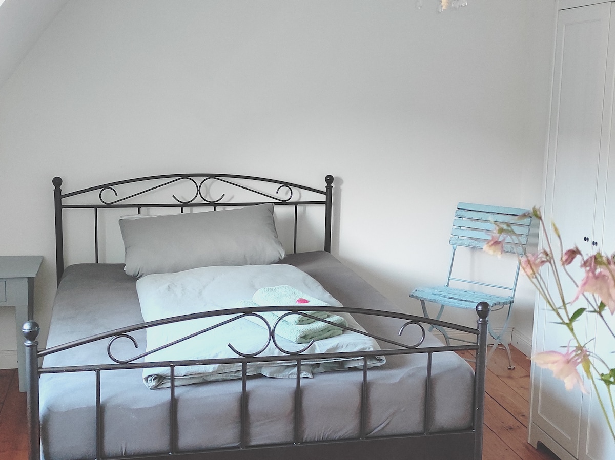 A spacious bed with a gray blanket and a decorative pillow is positioned centrally in the room. An accent chair in light blue is placed beside a minimalist bedside table, while a closet occupies the opposite wall. Soft natural light enhances the neutral color palette.