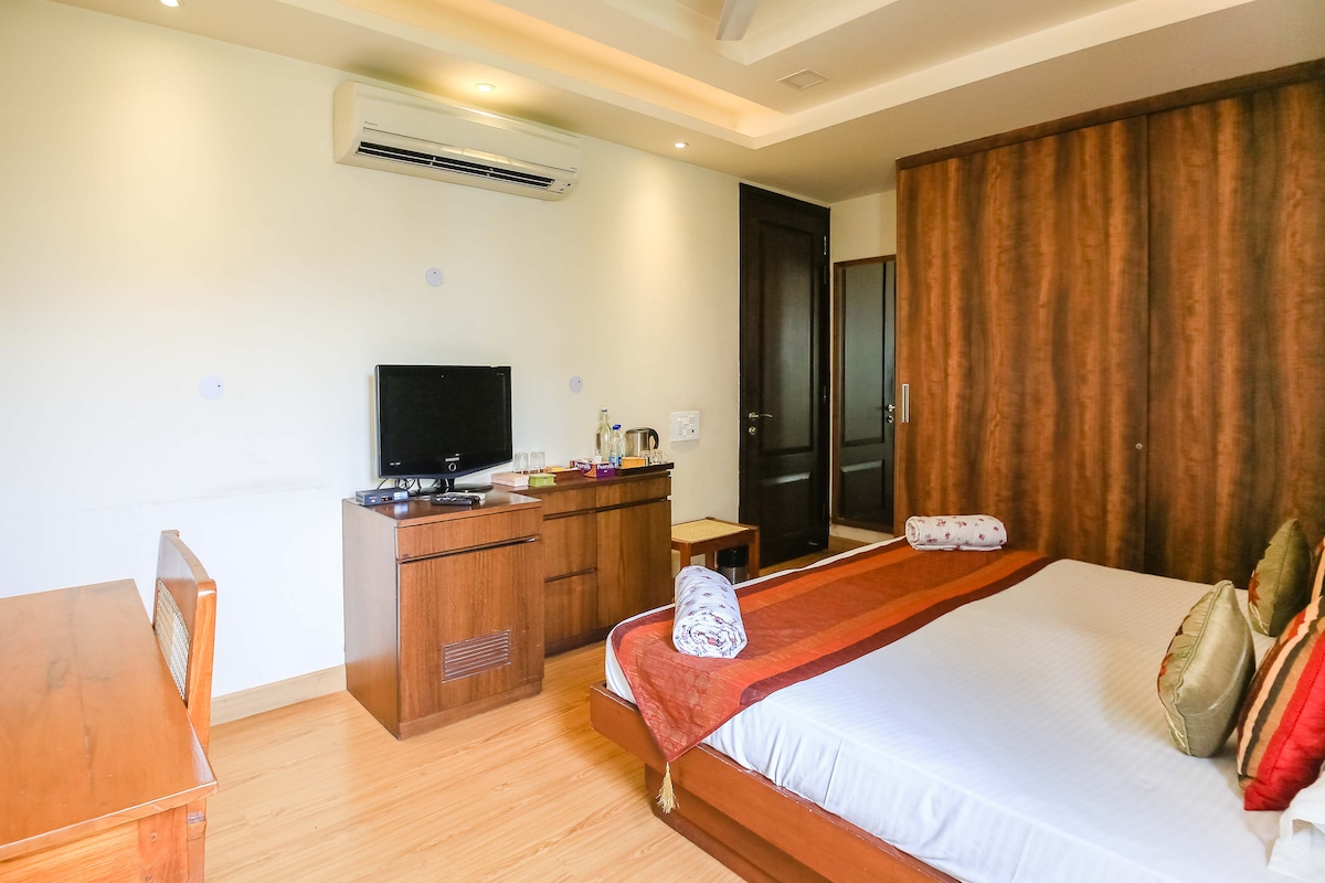 A well-appointed room features a spacious bed with decorative pillows. A wooden television cabinet holds a flat-screen TV and tea-making facilities. Natural light fills the area, and a large wardrobe with a dark wood finish complements the neutral walls. An air conditioning unit is mounted above.
