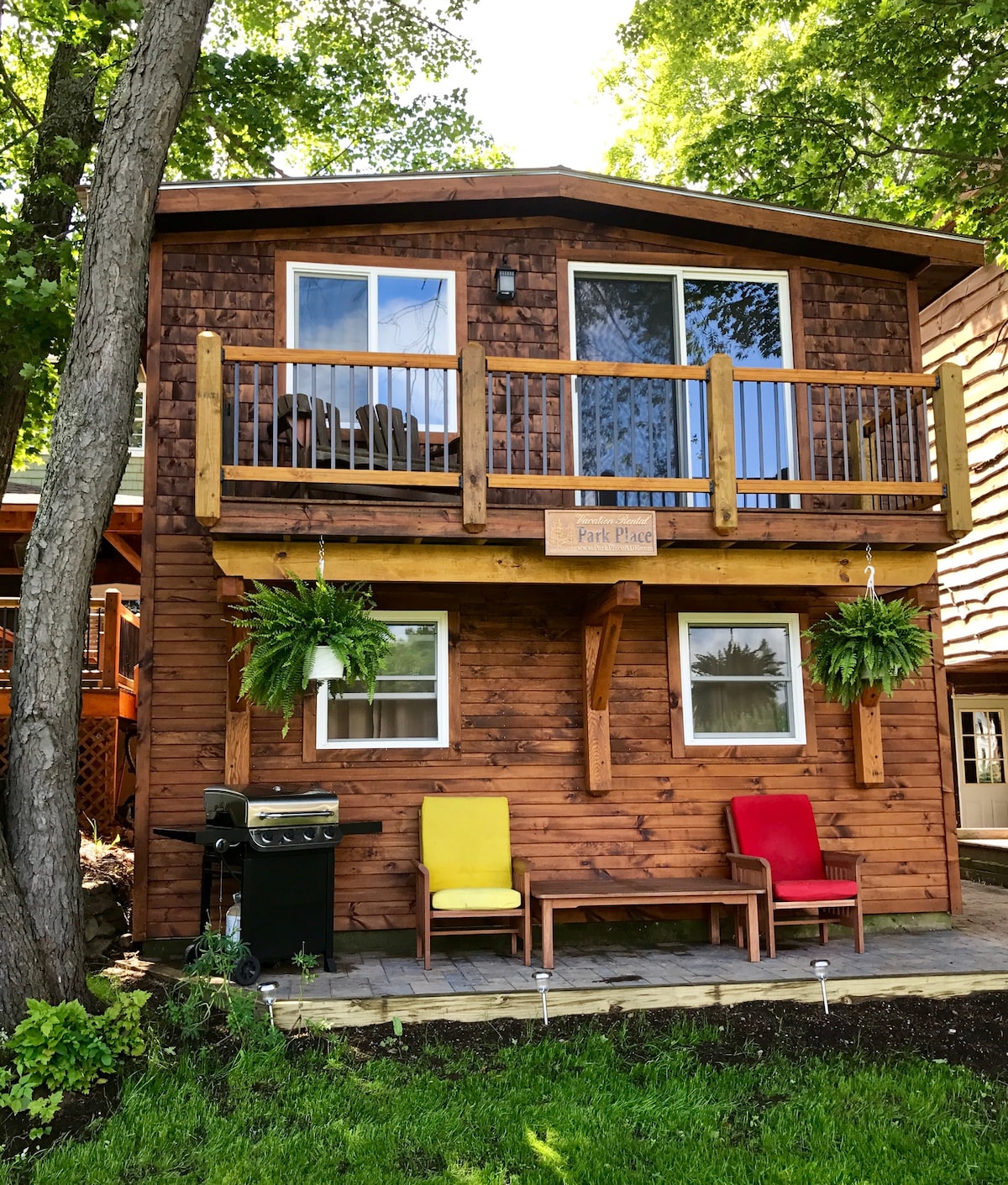 The exterior of the cottage is shown, featuring natural wood siding and a welcoming front porch. Two colorful chairs are placed on the patio, and a BBQ grill is available nearby. Lush greenery surrounds the space, creating a serene outdoor setting.