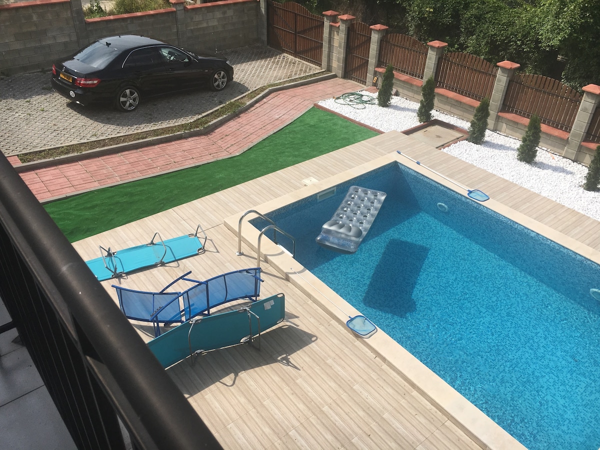 A spacious outdoor area features a clear swimming pool surrounded by wooden decking. Multiple blue loungers are positioned on one side, and a floating mat rests on the water's surface. A car is parked on the cobblestone driveway, and manicured shrubs line the property.