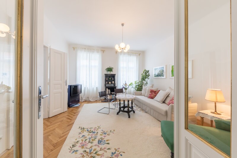 A spacious living room is visible, featuring large windows that allow natural light to brighten the space. A comfortable sofa and a decorative chair are arranged around a central coffee table. A patterned area rug adds warmth, complemented by plants and art on the walls.