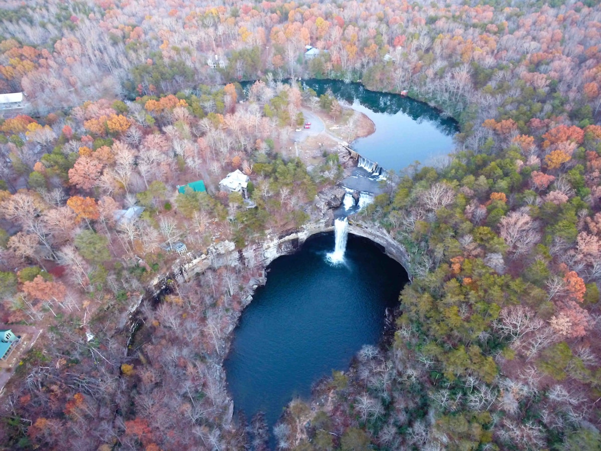 An aerial view captures a scenic landscape featuring a waterfall cascading into a dark blue pool. Surrounding the water are vibrant autumn-colored trees, with hints of green and brown. A small cabin is visible in the distance near the water's edge.